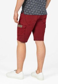 Red cotton cargo shorts with multiple pockets, stitched detailing, and a waistband label, paired with white sneakers and patterned shirt.