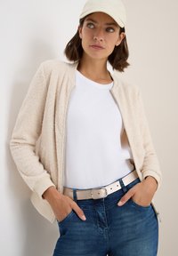 Beige fleece bomberjacka med ribbade manschetter, bärs över en vit t-shirt. Matchas med blå jeans och ett ljust bälte.