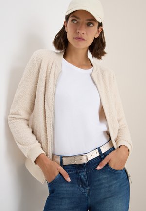Beige Fleece-Bomberjacke mit gerippten Bündchen, getragen über einem weißen T-Shirt. Kombiniert mit blauen Jeans und einem hellen Gürtel.