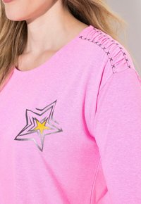 Felpa rosa con scollatura a barca, dettaglio arricciato in tessuto sulla spalla e una grafica di una stella nera e gialla sul petto.