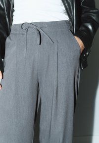 Grå skræddersyede bukser med bindebånd, med plisseringer og en blød tekstur, kombineret med en tætsiddende hvid top og en sort læderjakke.