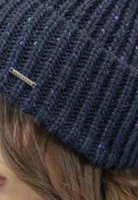 Sötétkék kötött sapka függőleges bordázással, világosabb kék foltokkal és ezüst logótáblával az oldalon. Puha tapintás és testhezálló forma.