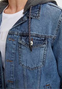 Giubbotto di denim blu chiaro con cappuccio grigio. Le caratteristiche includono due tasche sul petto, cuciture in rame e un cordino a contrasto.