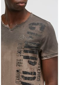 V-neck t-shirt in muted brown, featuring distressed edges, screen-printed black text, and subtle logo detailing on the front.