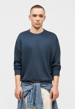 Jonge persoon met kort haar, die een donkerblauwe trui en een blauw geruit overhemd rond de taille geknoopt draagt, staand tegen een effen achtergrond.