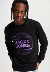 Svart hettegenser med lilla grafisk trykk som viser "JACK & JONES CORE". Laget av bomull, rund hals, ribbestrikkede ermer. Casual passform.