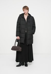 Mujer vestida con un abrigo largo de plumas negro, falda negra, botas negras, sosteniendo un bolso oscuro, de pie contra un fondo blanco.