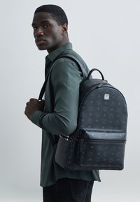 Mochila negra con textura y patrón de monograma, compartimento principal grande, bolsillo frontal con cremallera y detalles con tachuelas. Llevada sobre el hombro izquierdo.