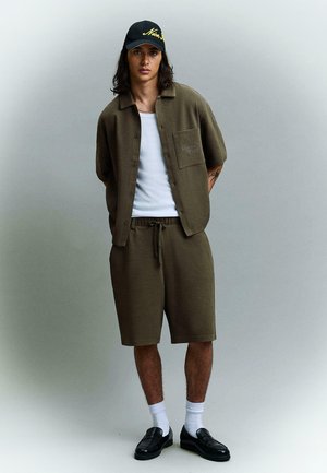 Persoană tânără purtând cămașă verde măsliniu cu mâneci scurte și pantaloni scurți asortați, maieu alb, șapcă neagră, șosete albe și mocasini negri, stând relaxată.