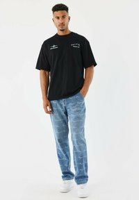 Joven de pie con una mano en el bolsillo, vistiendo una camiseta negra con gráfico, jeans azules con estampado de letra blanca y zapatillas blancas.