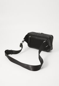 Black crossbody bag made of textured material, featuring a zip closure, mesh panel, and adjustable strap. Compact rectangular shape.