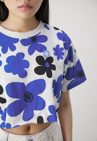 Camiseta recortada de algodón blanco con un patrón floral en azul y negro. Presenta un pequeño logo con una carita sonriente, mangas cortas y un escote redondo.