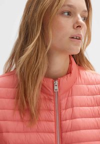 Veste en quilté légère de couleur corail, avec un col haut et une fermeture éclair centrale. Texture lisse avec un léger éclat.