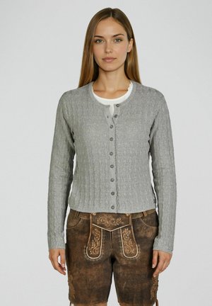 Jeune femme portant un cardigan gris à torsades par-dessus un haut blanc et un short en cuir traditionnel brodé marron, debout devant un fond uni.