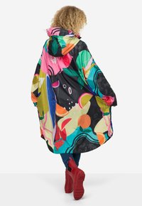 Färgglad vindjacka med huva, prydd med djärva blommiga och abstrakta mönster på en svart bakgrund, tillverkad av lätt material. Röda skor synliga.