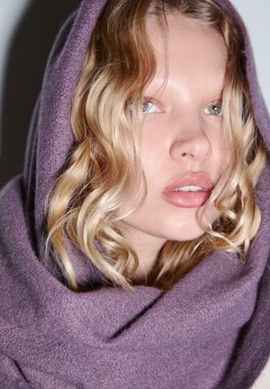 Sciarpa di lana viola drappeggiata attorno al viso, texture morbida, che mette in risalto i capelli biondi lucidi con onde morbide e labbra lisce dai toni neutri.