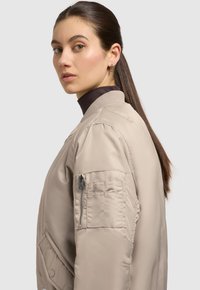 Beige Bomberjacke mit einem gerippten Kragen, seitlichen Taschen und einer Reißverschlusstasche am Ärmel. Glatte Textur mit minimalen Nähdetails.