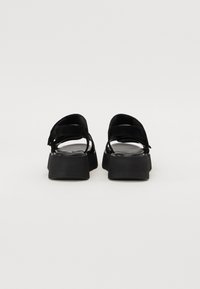 Musta pläkkiläiset sandaalit, joissa on kaksi säädettävää hihnaa. Valmistettu sileästä nahkamaisesta materiaalista. Ominaisuuksiin kuuluu leveä, paksu pohja ja minimalistinen muotoilu.