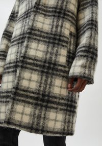 Abrigo de lana a cuadros en crema, negro y gris con una textura suave, que presenta un patrón de cuadros y mangas largas. Destaca por su corte exagerado.