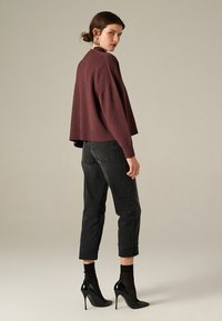 Maglione corto bordeaux con orlo a coste, abbinato a jeans scuri alta vita. Il modello indossa scarpe nere con tacco alto e calze trasparenti, posando di schiena.