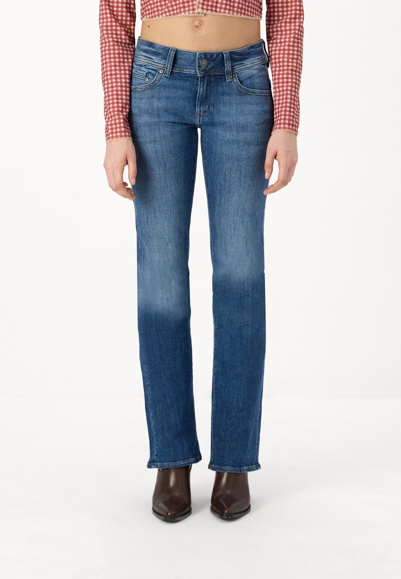 Femme portant un jean droit bleu taille mi-haute, des bottines en cuir marron pointues et un haut court à manches longues à carreaux rouge et blanc.