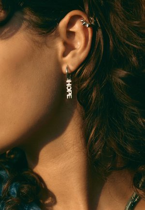 Nahaufnahme eines Ohrs einer Person mit lockigem Haar, die einen Creolen-Ohrring trägt, der das Wort "HOPE" bildet, sowie eine mit Spitzen versehene Manschette am oberen Ohr.