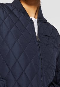 Gros plan d'une personne portant une veste matelassée bleu marine avec une fermeture éclair à l'avant et un col côtelé noir par-dessus une chemise blanche.