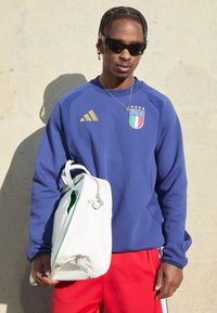 Plavi dukserica dugih rukava s zlatnim detaljima i logom Italije, crvene hlače, drži bijelu torbu s zelenim obodom i srebrnim zatvaračem.