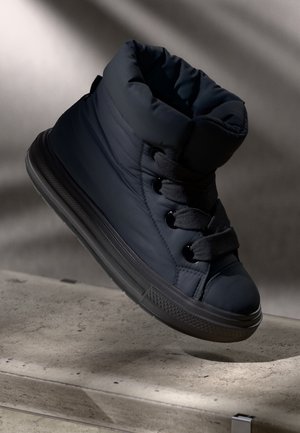 Zwarte gevoerde hoge sneaker met dikke zool en brede veters, getoond boven een gestructureerd betonnen oppervlak met zacht licht.