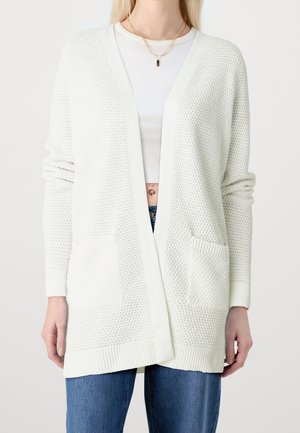 Femme portant un cardigan blanc texturé ouvert sur un haut blanc court, un jean bleu et des colliers en or superposés, debout devant un fond blanc.