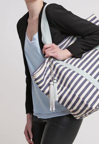 Bolsa de tela a rayas en colores marino y crema, con una correa de color azul claro, detalle de cadena y acentos de borlas. Textura suave.