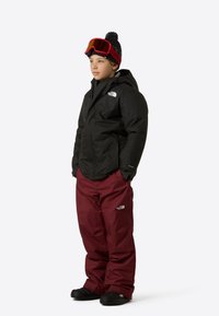 Giacca impermeabile nera con cappuccio, pantaloni da neve color vinaccia, stivali invernali neri e occhiali da sci rossi con un cappello a pompon nero e rosso.