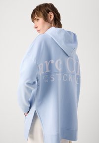 Felpa con cappuccio di colore blu chiaro con uno spacco laterale, caratterizzata da un testo bianco che recita "est. in STOCKHOLM." Tessuto morbido con una vestibilità rilassata.