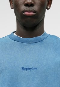 Ανοιχτού μπλε φούτερ με ριγέ γιακά, μαλακή βαμβακερή υφή και κεντημένο λογότυπο "Replay Inc." σε σκούρο μπλε.
