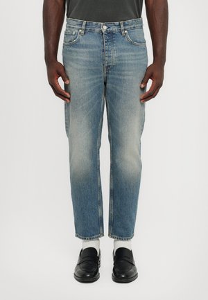 Blugi de denim albastru deschis cu croială dreaptă, aspect decolorat și design cu cinci buzunare. Poartă cu mocasini negri și șosete albe.