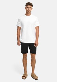 Weißes Baumwoll-T-Shirt, kurze Ärmel, runder Ausschnitt, kombiniert mit schwarzen Shorts, umgeschlagenen Säumen und braunen Sandalen. Einfaches, lässiges Outfit.