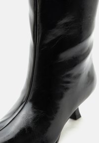Μαύρο δερμάτινο μπότες έως τη μέση της γάμπας με διακριτική λάμψη και χαμηλό, κεκλιμένο τακούνι σε λευκό φόντο.