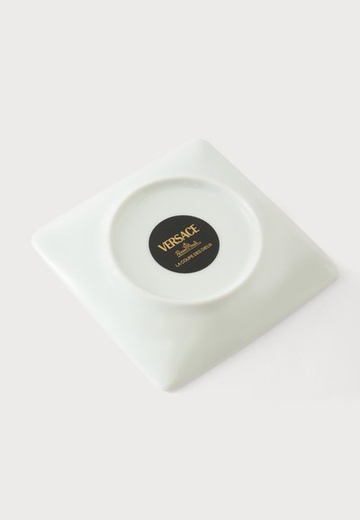 Assiette carrée en céramique blanche avec une indentation circulaire et une étiquette Versace noire en lettres dorées au centre.
