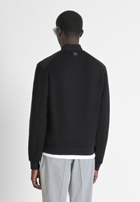 Felpa nera con un design sportivo, polsini a coste e un logo sottile nel colletto. Indossata sopra una camicia bianca e abbinata a pantaloni grigio chiaro.