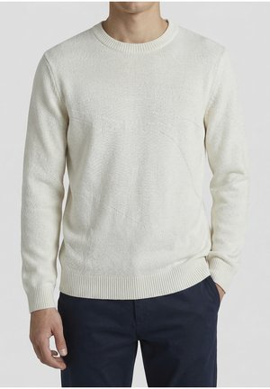 Man draagt een crèmekleurige gebreide trui met een gestructureerd patroon en geribbelde manchetten en zoom, gecombineerd met donkerblauwe broek, staat met één hand in de broekzak.