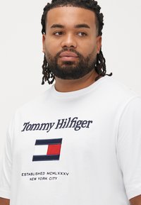 White cotton T-shirt with a crew neck, featuring embroidered "Tommy Hilfiger" logo and rectangular flag graphic in red, white, and blue.