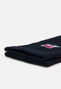 Jūras spēku zili riboti adīti zeķes, kurām ir neliels izšūts logo ar sarkaniem un baltiem akcentiem. Salocītas, lai parādītu tekstūru un dizaina detaļas.