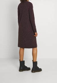 Person klädd i en knälång mörklila långärmad klänning och svarta snörade ankelstövlar, står mot en enkel vit bakgrund.