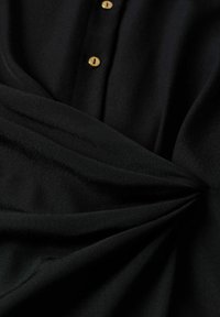 Tissu noir avec deux boutons beiges visibles et un détail froncé torsadé à la taille, mettant en valeur la texture et les coutures.