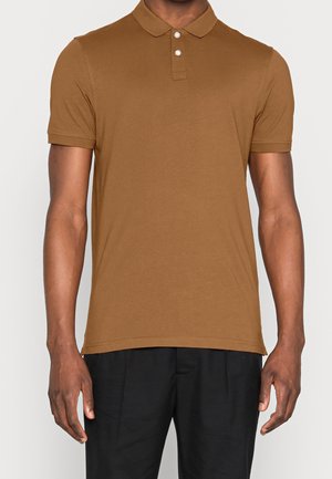 Homme portant un polo marron à manches courtes avec deux boutons blancs et un pantalon noir, debout devant un fond uni.