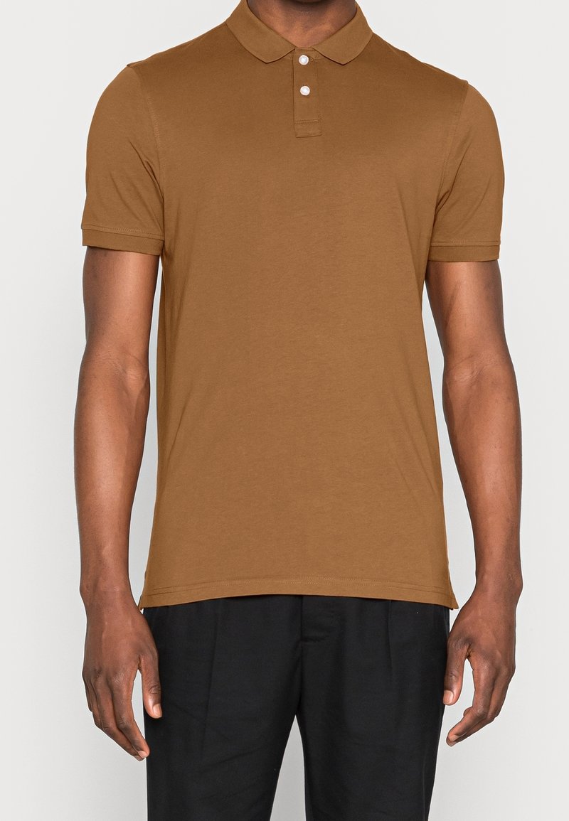 Homme portant un polo marron à manches courtes avec deux boutons blancs et un pantalon noir, debout devant un fond uni.