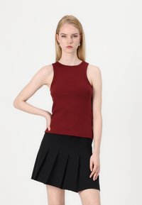 Burgunderfarget ribbet tanktopp med rund hals, uten ermer, kombinert med et svart plissert skjørt. Glatt tekstur, tettsittende silhuett.