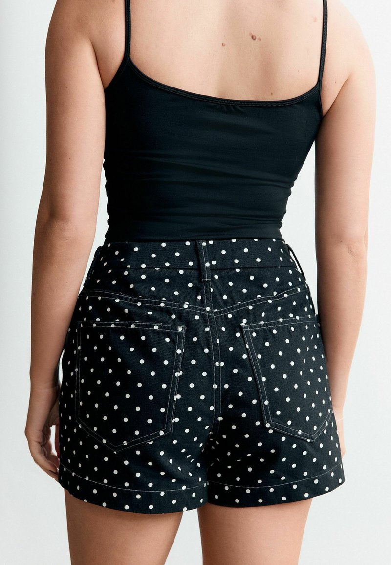 Femme portant un haut noir à fines bretelles et un short taille haute noir à pois blancs, vue de dos sur un fond blanc.