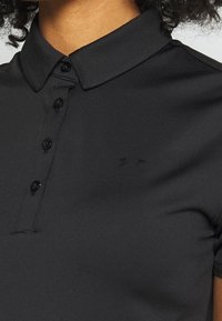Gros plan sur un polo noir avec des boutons et un petit logo Under Armour discret sur la poitrine.