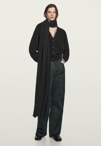 Czarny dzianinowy kardigan z guzikami z przodu, w połączeniu z ciemnymi dżinsami o szerokich nogawkach i długim czarnym szalikiem owiniętym wokół szyi. Widoczne detale teksturowanej tkaniny.